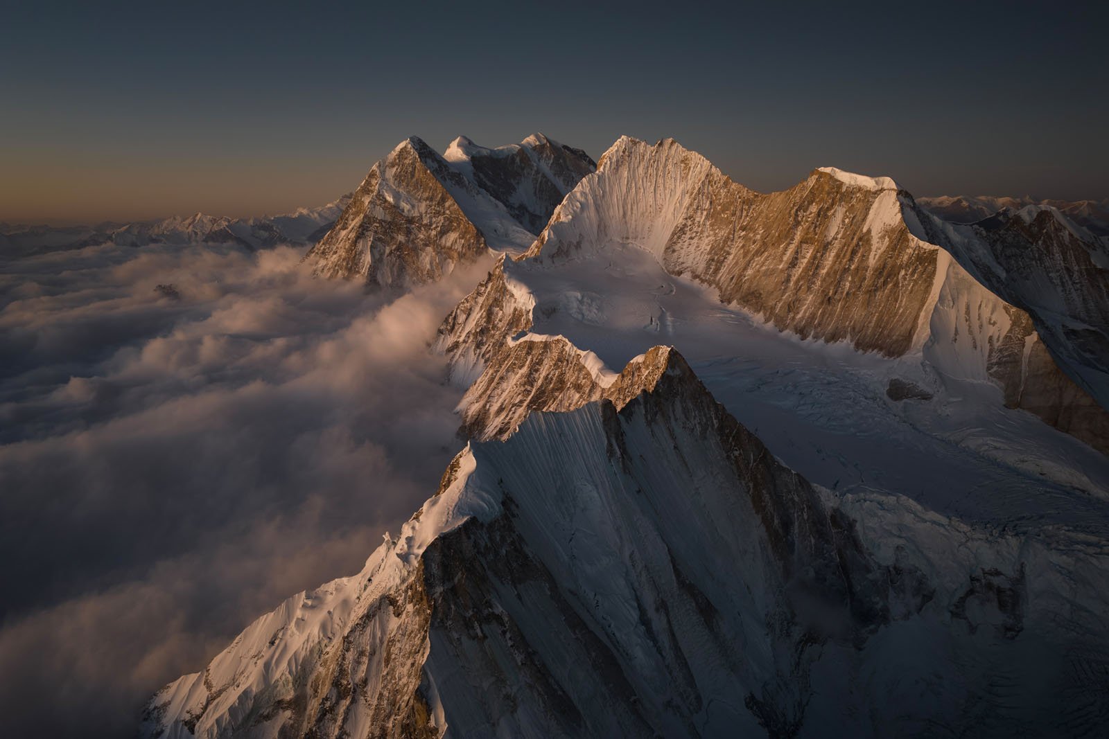 Snow-capped mountain peaks rise above a sea of clouds at sunrise, with warm golden light illuminating the sharp ridges and casting dramatic shadows over the landscape.