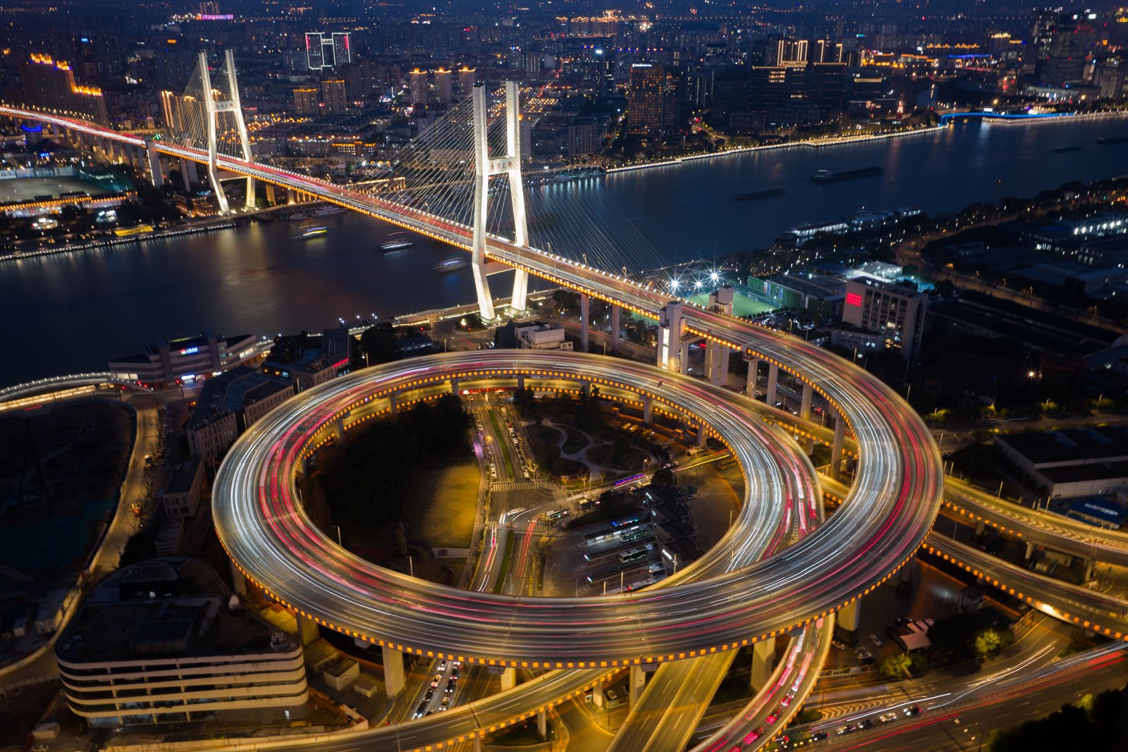 Aerial view of a large illuminated bridge crossing a river at night, with a glowing circular highway interchange in the foreground and city buildings in the background.