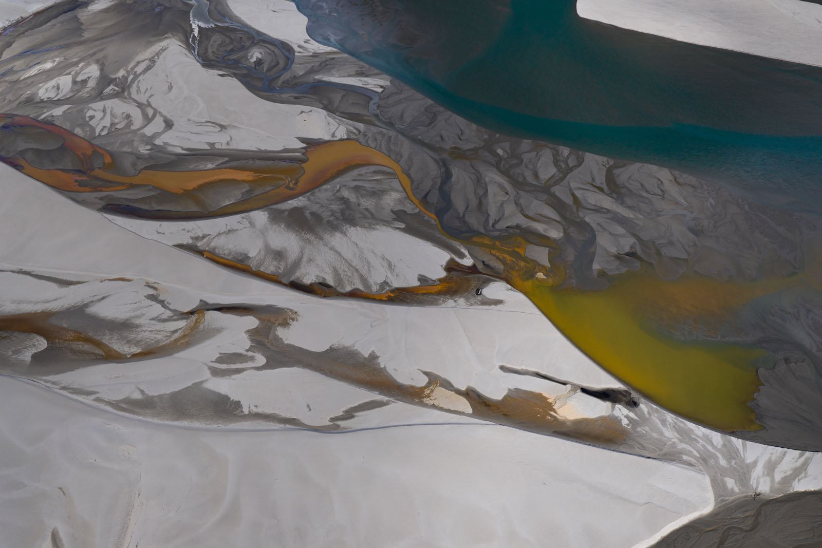 Aerial view of a river delta with winding channels of blue and yellow water cutting through white and gray sand, creating abstract, painterly patterns.