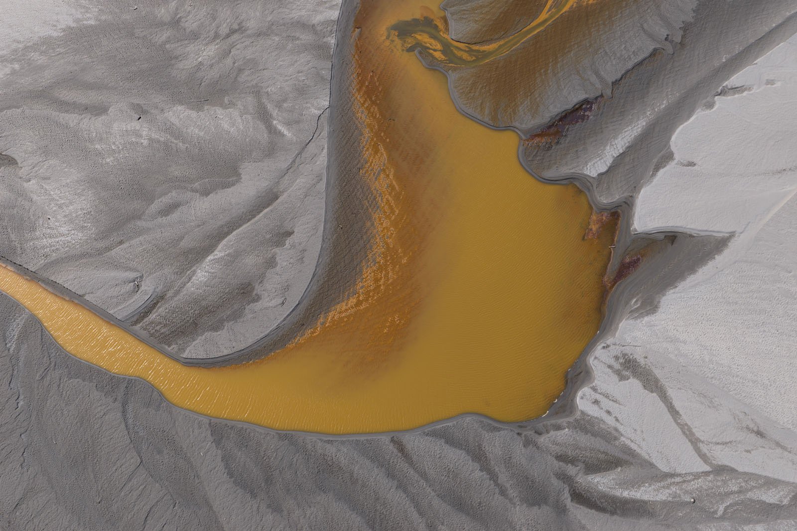 Aerial view of a winding river or stream with striking orange water flowing through a landscape of gray and white sand or sediment, creating an abstract, textured pattern.