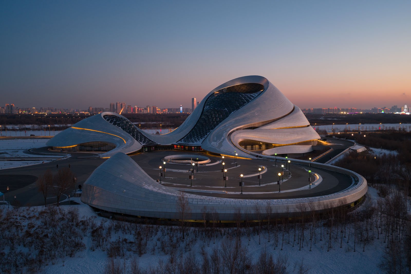 A futuristic building with smooth, curved lines and a glass dome sits in a snowy landscape at dusk, with city lights and a skyline visible in the background.