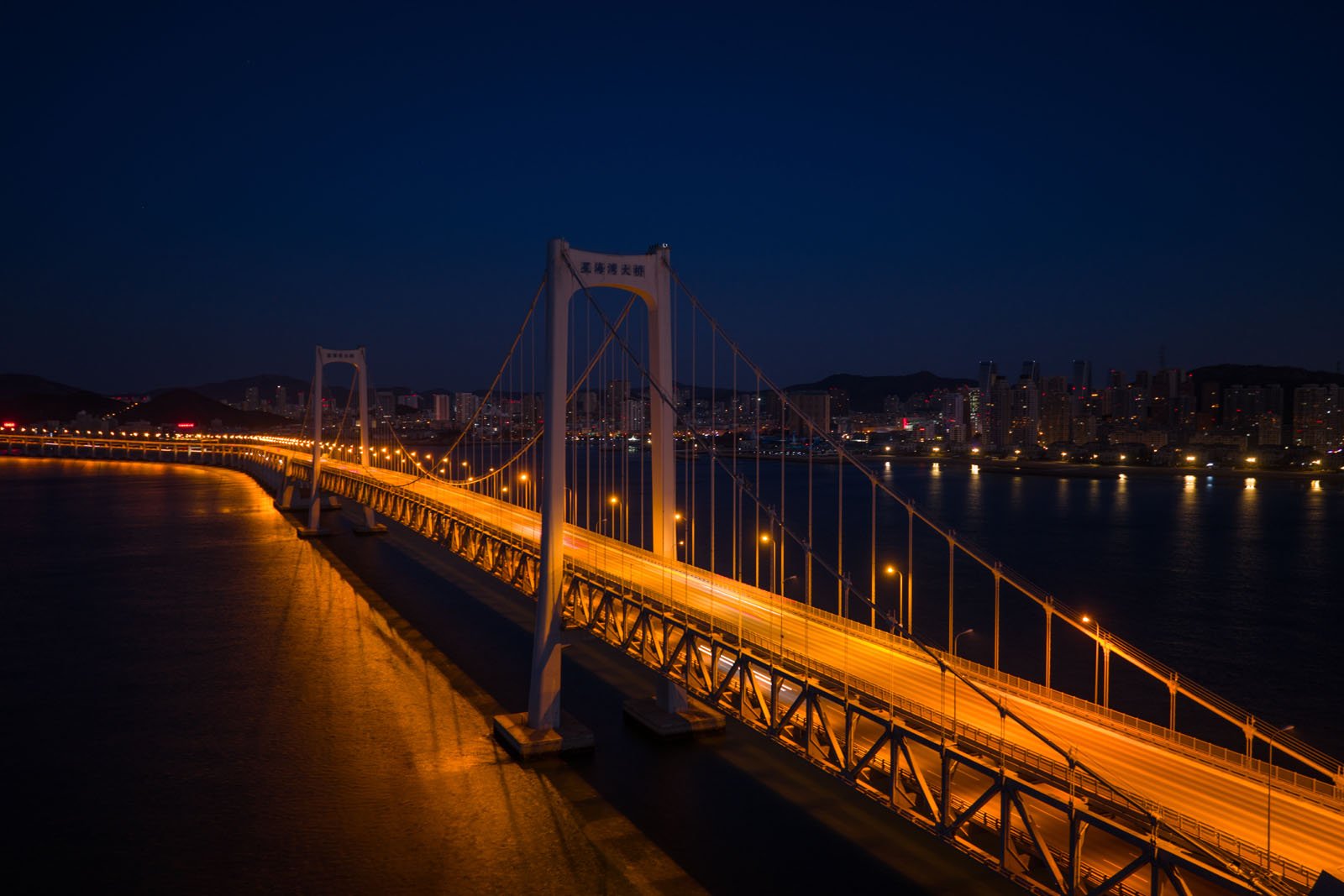 Night view of a brightly lit suspension bridge spanning across water, with illuminated city buildings in the background and dark hills on the horizon.