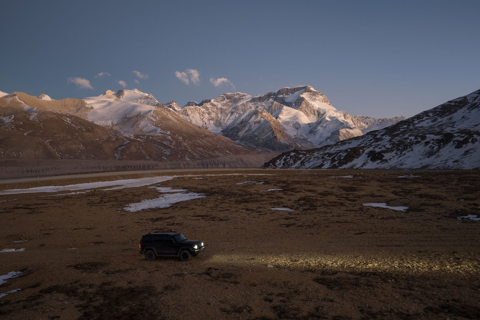 A black SUV with headlights on drives across a barren, rocky landscape at dusk, with snow-capped mountains rising majestically in the background under a clear sky.