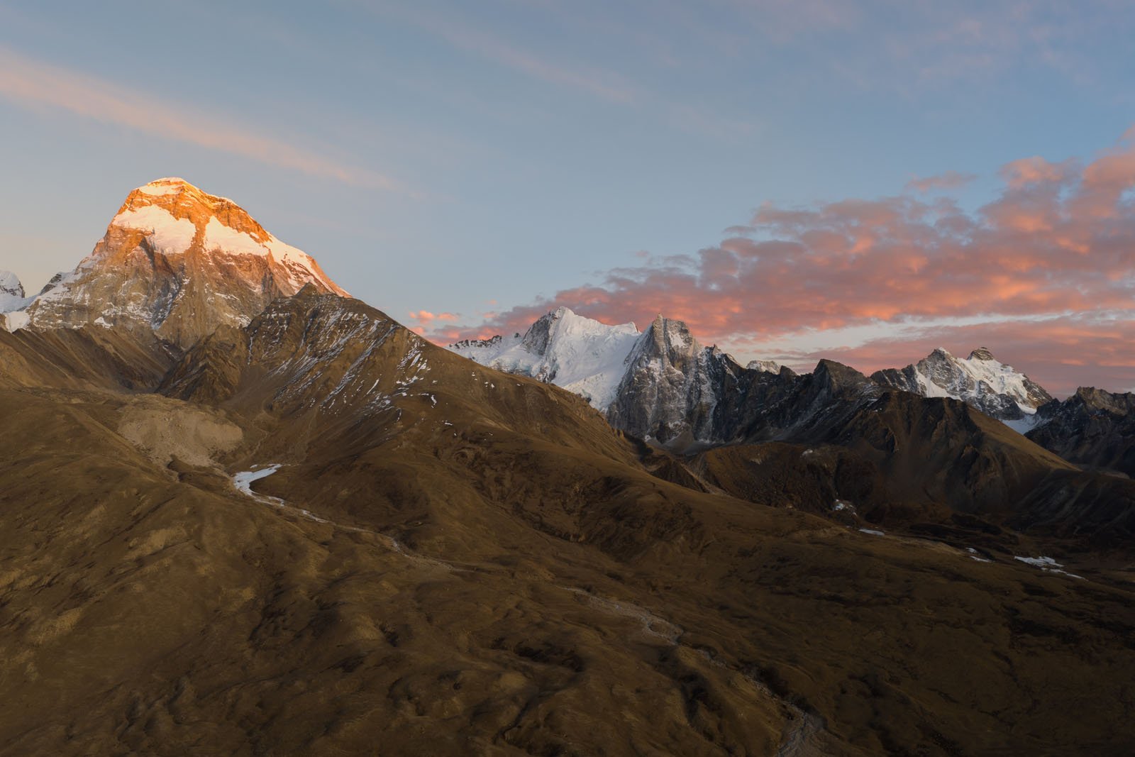 A mountain range at sunrise, with the tallest peak bathed in golden light, surrounded by rugged brown slopes and patches of snow under a sky with pink clouds.