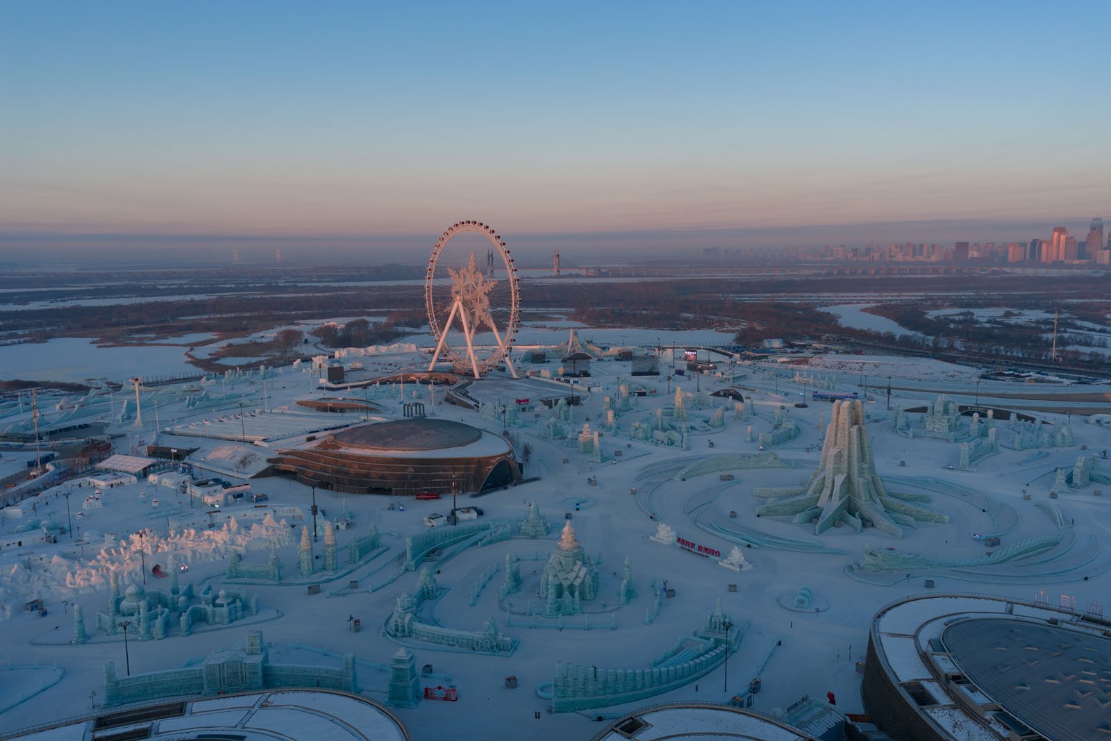 Aerial view of a large ice and snow festival with intricate ice sculptures, a Ferris wheel, and surrounding buildings at dawn; a river and city skyline appear in the distance under a clear sky.