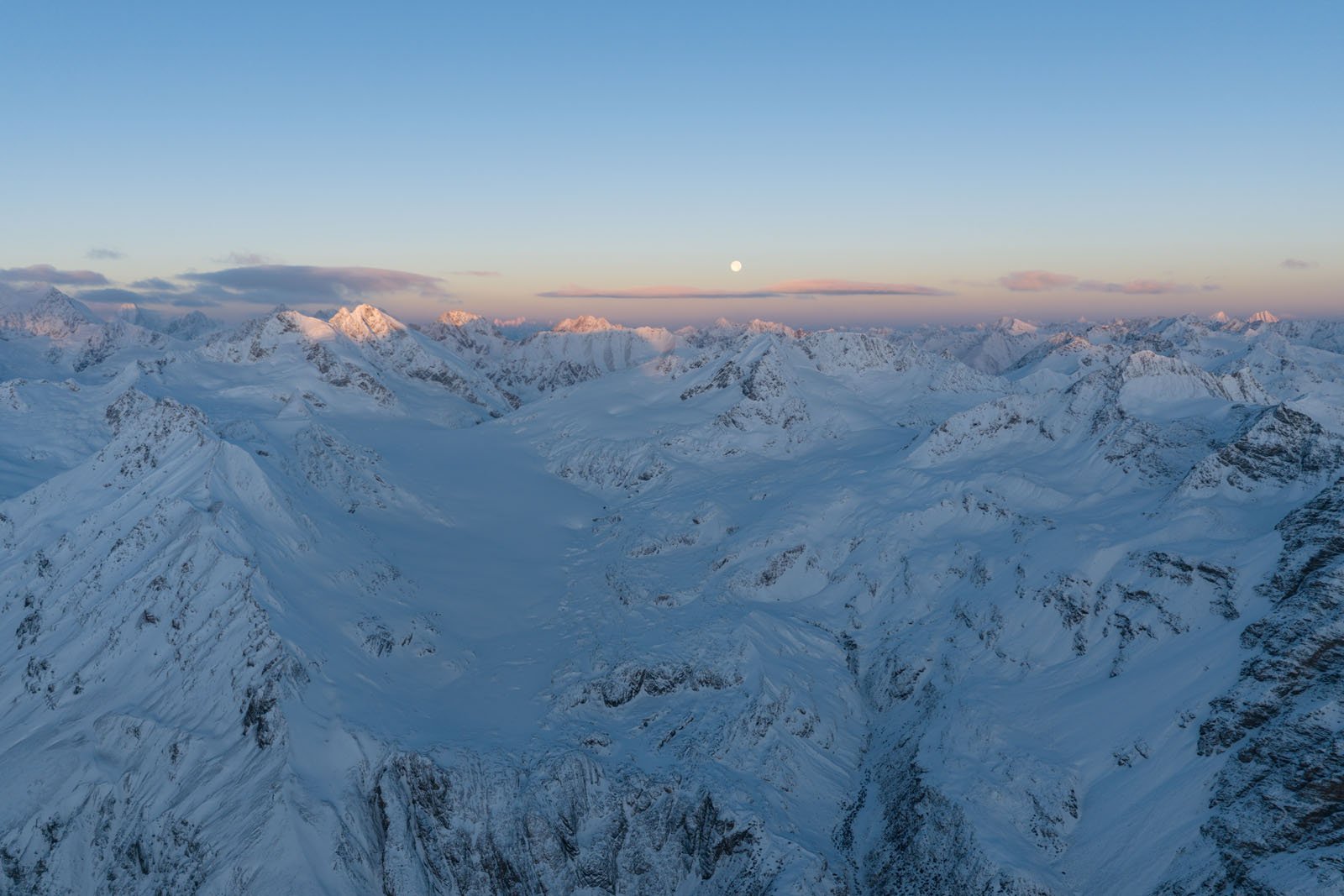 Aerial view of snow-covered mountain peaks at sunrise or sunset, with a full moon visible in the clear sky above the rugged, icy landscape.
