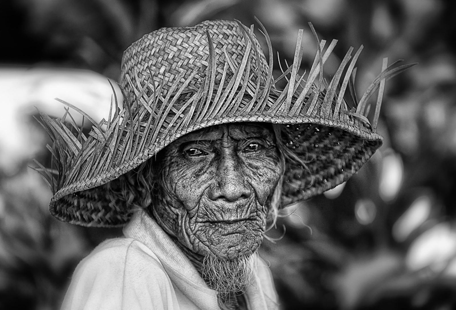 An elderly person with deep facial wrinkles wears a woven straw hat with spiky edges. The black and white photo highlights the texture of their skin and hat, with a blurred natural background.