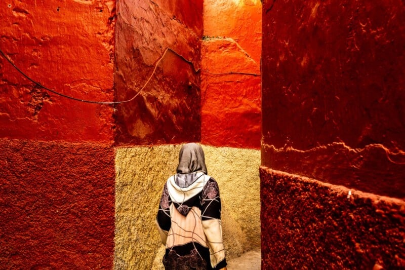 A person wearing a hooded jacket and scarf walks through a narrow alleyway with vibrant red and yellow textured walls.