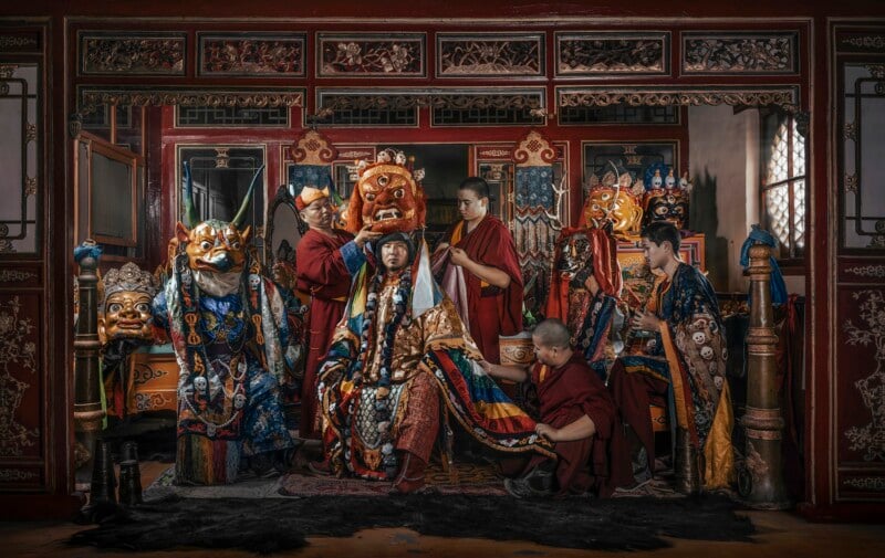 A group of people in ornate traditional costumes and masks prepare for a performance or ritual inside an elaborately decorated room with red and gold details. Some adjust a central figure's costume and mask.