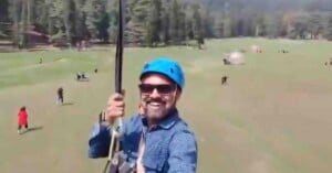 A man wearing a blue helmet and sunglasses smiles while ziplining over a grassy open field with trees and people in the background.