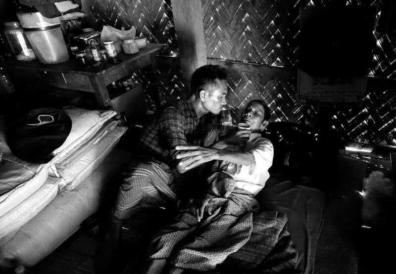 A man sits on a bed, gently supporting and feeding another man who is lying down and appears unwell, inside a dimly lit, woven bamboo hut with basic supplies on a shelf nearby.