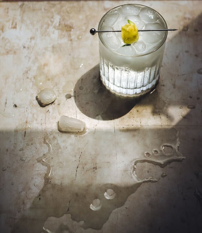 A glass of ice-filled cocktail garnished with a lemon twist on a skewer sits on a wet surface, surrounded by melted ice and water droplets in dramatic sunlight and shadow.
