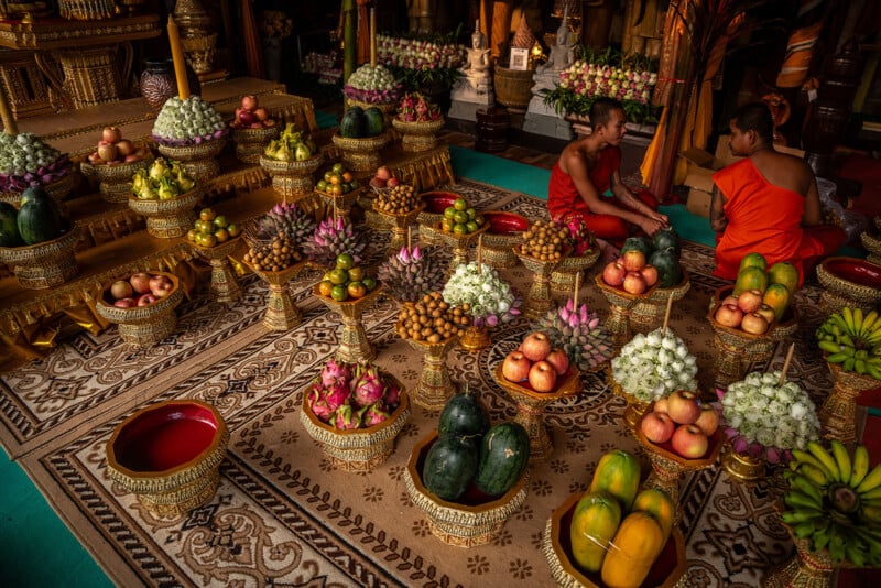 Two monks in orange robes sit among ornate golden trays filled with various fruits and flowers arranged on patterned carpets in a temple setting, creating a vibrant and colorful display.