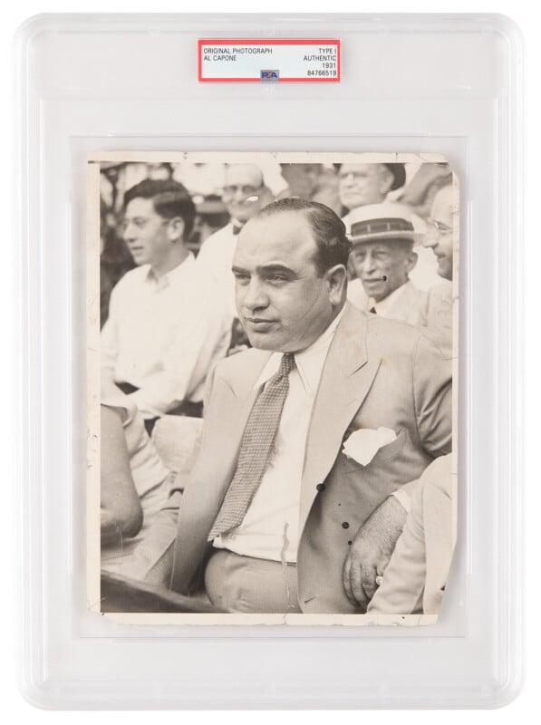 Black-and-white photo of a man in a suit and tie sitting among a crowd, encased in a graded display with a label reading "Original Photograph Al Capone." The man appears calm and is looking to the side.