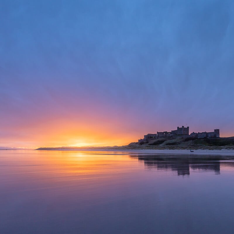 A castle sits on a hill overlooking a calm beach at sunrise or sunset, with vibrant orange and purple hues reflected on the wet sand and water. The sky is partly cloudy, deepening the scene’s colors.