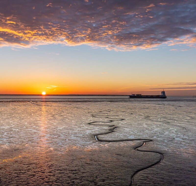 A cargo ship sails across a calm sea at sunset, with the sun low on the horizon, streaks of clouds above, and winding water channels visible on the wet, reflective shoreline in the foreground.