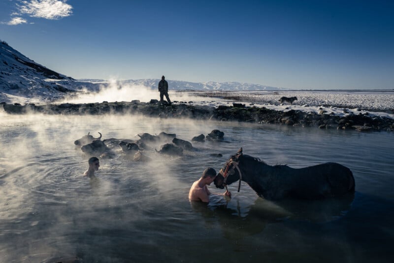 Two men and a horse bathe in a steaming hot spring on a winter day, with snow-covered ground, mountains in the background, and another person and animals standing nearby. Steam rises from the water into the cold air.