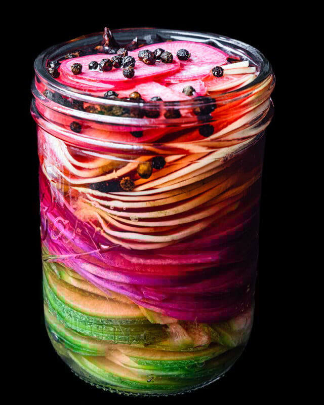 A glass jar filled with thinly sliced, colorful vegetables including cucumbers, red onions, and radishes, layered neatly and topped with whole black peppercorns, all in clear pickling liquid against a black background.