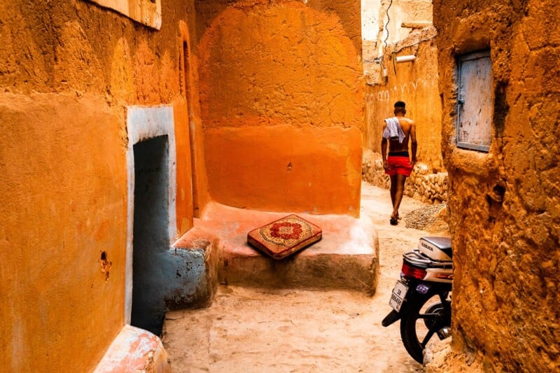 A person walks through a narrow, sunlit alley with orange and clay-colored walls, a patterned rug on a step, and a parked motorcycle in the foreground. The scene feels warm and vibrant.