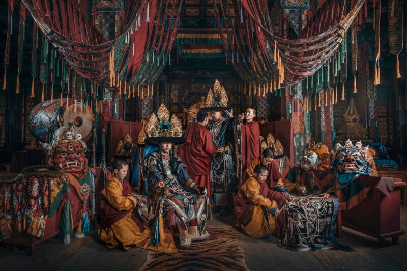 A group of monks and young boys in traditional Tibetan attire sit and stand in a richly decorated temple, surrounded by ornate masks, drums, and ceremonial objects, under hanging red and gold banners.