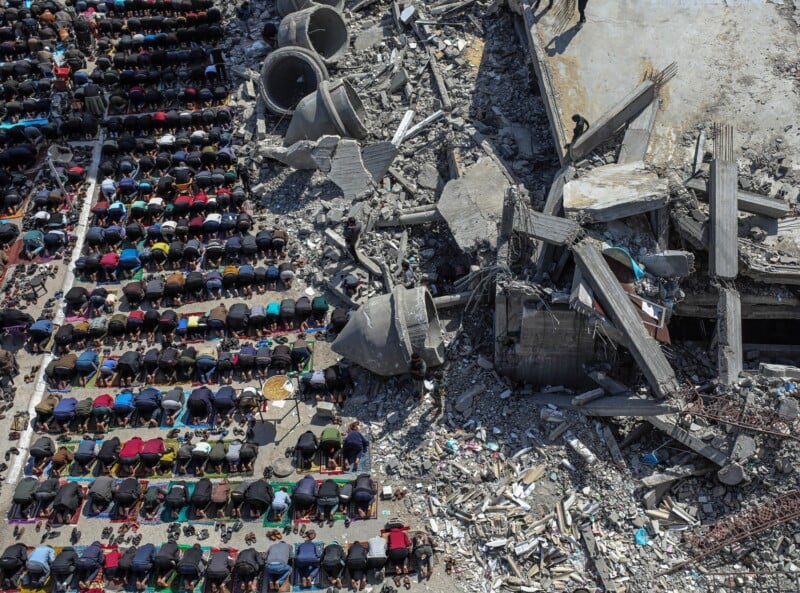 A large group of people pray in neat rows on the ground next to the rubble and debris of destroyed buildings, highlighting a stark contrast between the act of worship and surrounding devastation.