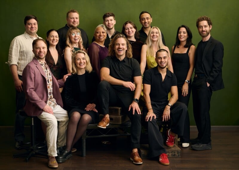 A group of fifteen people, dressed in semi-formal attire, pose together and smile against a green background. Some are sitting while others stand behind them, creating a friendly and cheerful atmosphere.