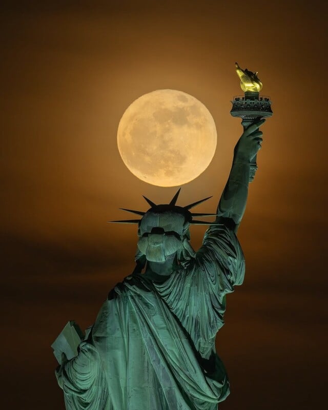 The Statue of Liberty is seen from behind at night, holding up her torch, with a large full moon glowing brightly in the cloudy sky directly above the flame.