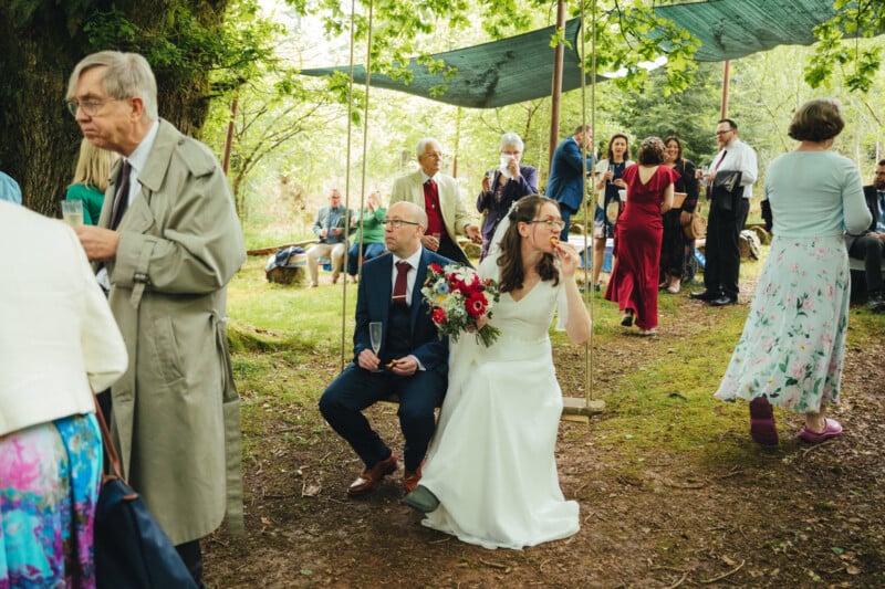 A bride in a white dress and groom in a suit sit on a wooden swing at an outdoor wedding, surrounded by guests socializing under a canopy of trees and green tarps. The bride holds a bouquet and laughs.