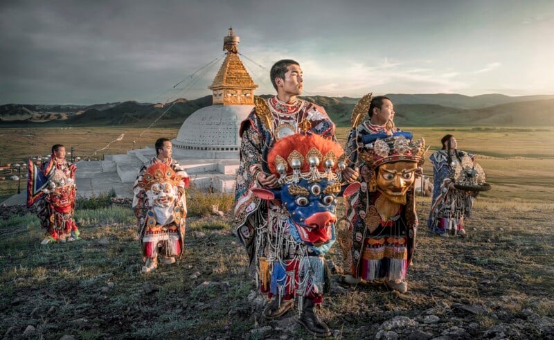 Five people in ornate, colorful traditional costumes and masks stand on a grassy hill near a large white stupa with golden details, surrounded by vast open landscape and distant mountains at sunset.