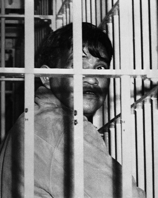 A man with short dark hair sits behind metal jail bars, looking upward. The image is black and white, and shadows partially obscure his face.