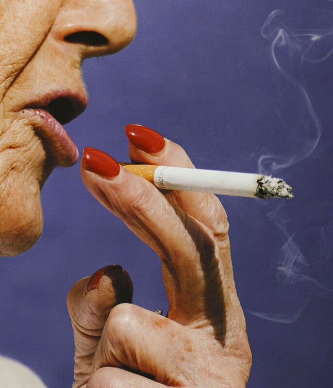 Close-up of an older person with red lipstick and red-painted nails holding a lit cigarette near their mouth, with visible smoke against a purple background.