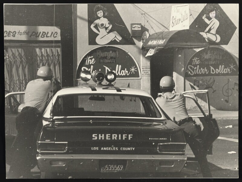Two sheriff deputies in helmets take cover behind a Los Angeles County sheriff car, aiming guns at a building with images of women and signs reading "The Silver Dollar." The scene appears tense and urban.