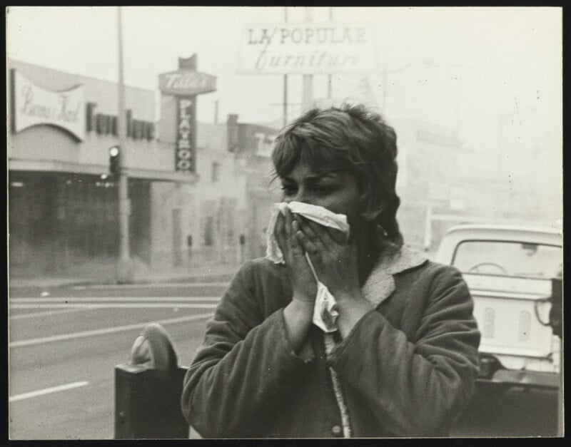 A woman stands on a city street, covering her nose and mouth with a tissue. The background is hazy with smoke or smog, and storefront signs and a parked car are visible behind her.