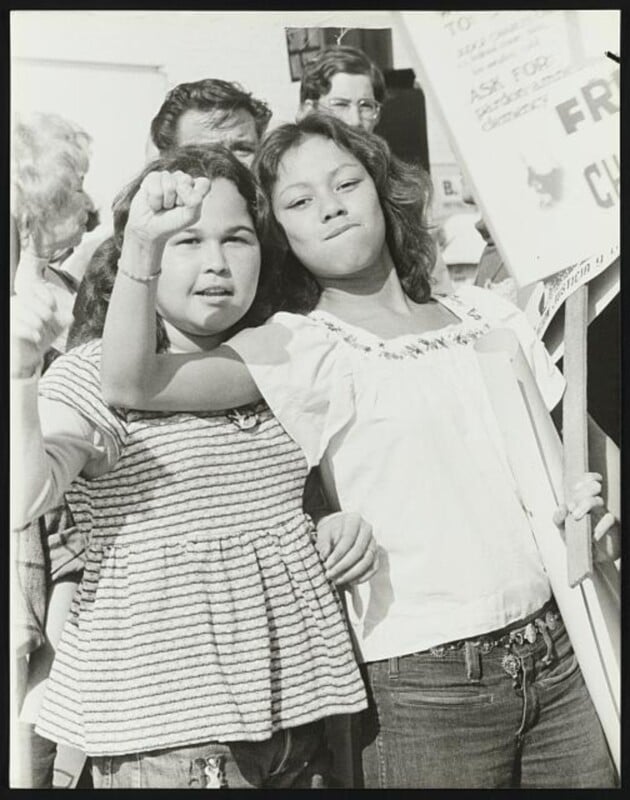Two young women stand side by side in a crowd at a protest, one raising her fist and the other holding a sign. Both look determined and confident. Other people and protest signs are visible in the background.