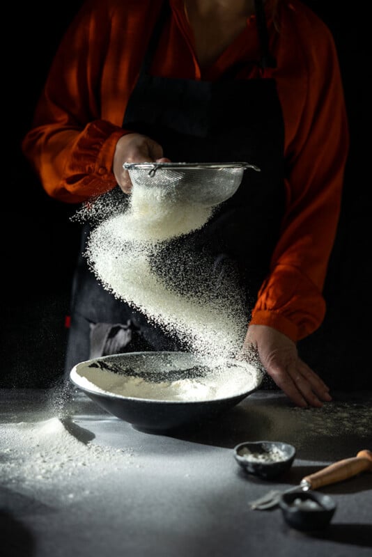 A person in an orange shirt and black apron sifts flour into a bowl, with the flour forming a swirling pattern in mid-air. The scene is set against a dark background with kitchen utensils nearby.