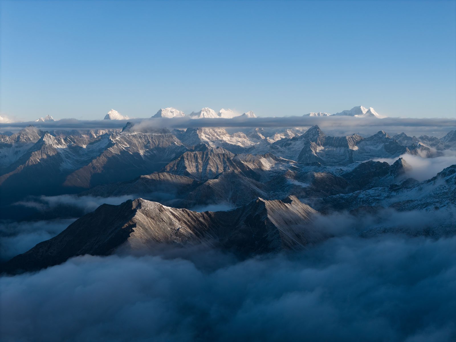 A panoramic view of snow-covered mountain peaks rising above clouds, with clear blue sky and a layer of mist partially covering the lower slopes.