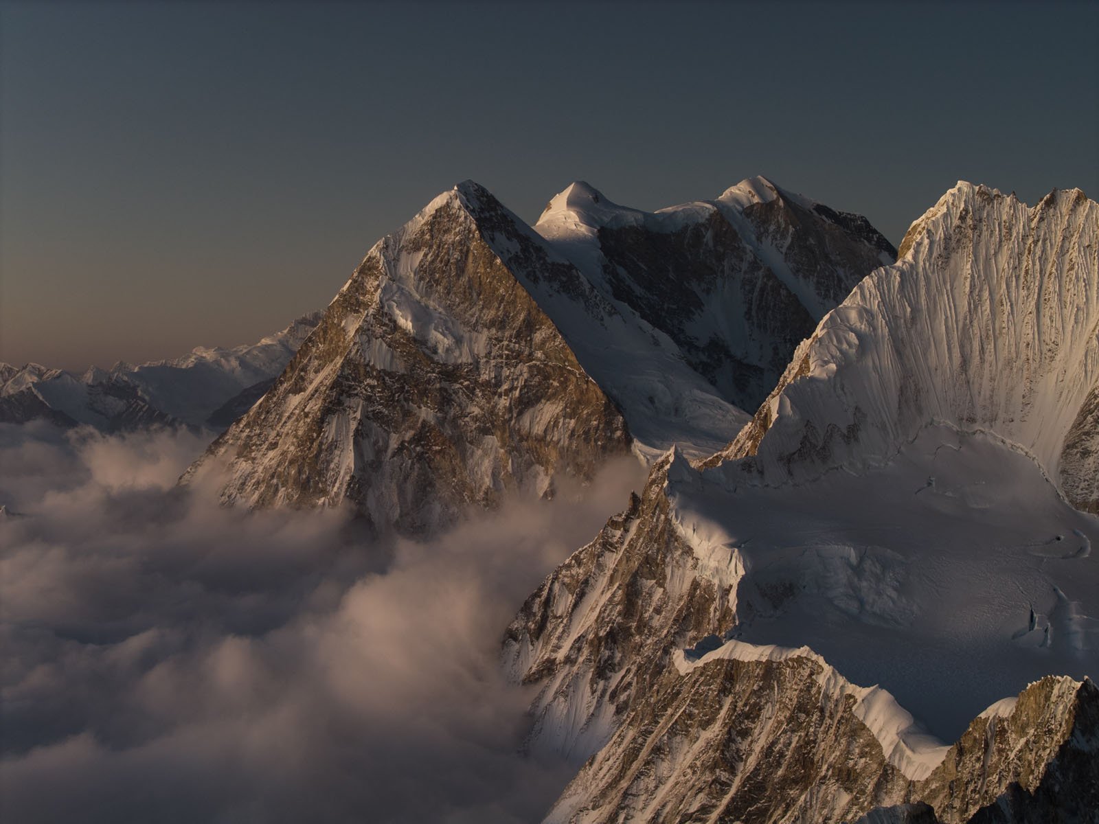 A dramatic view of snow-covered mountain peaks rising above a layer of clouds at sunset, with soft golden light illuminating the rugged slopes and ridges.