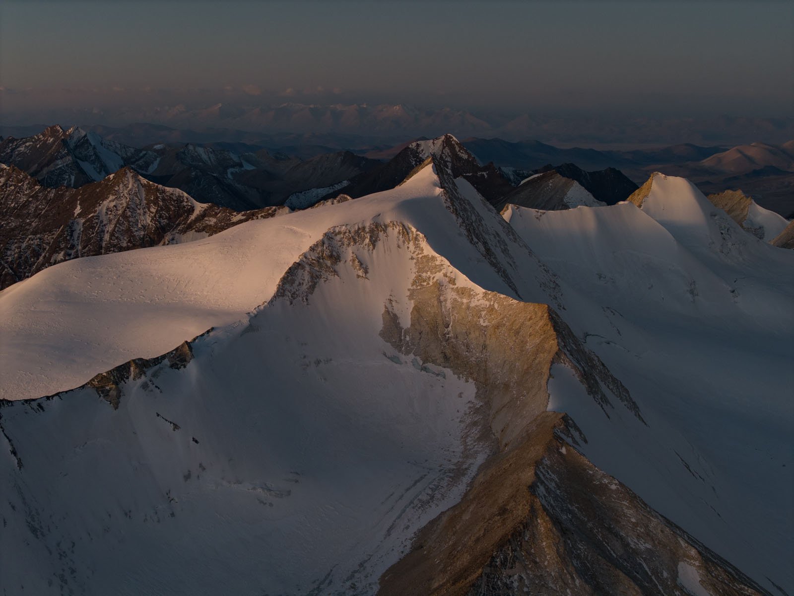 Snow-covered mountain peaks bathed in soft, golden light at sunrise or sunset, with rugged ridges and distant mountains fading into the hazy background.