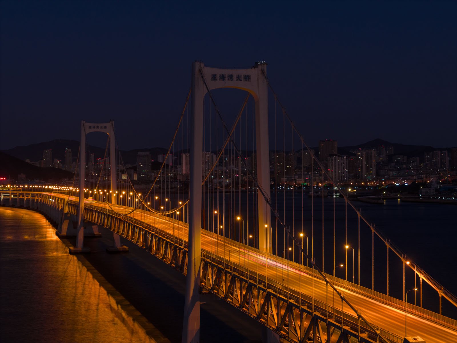 A suspension bridge illuminated by streetlights stretches across water at night, with a city skyline and buildings visible in the background under a dark sky.
