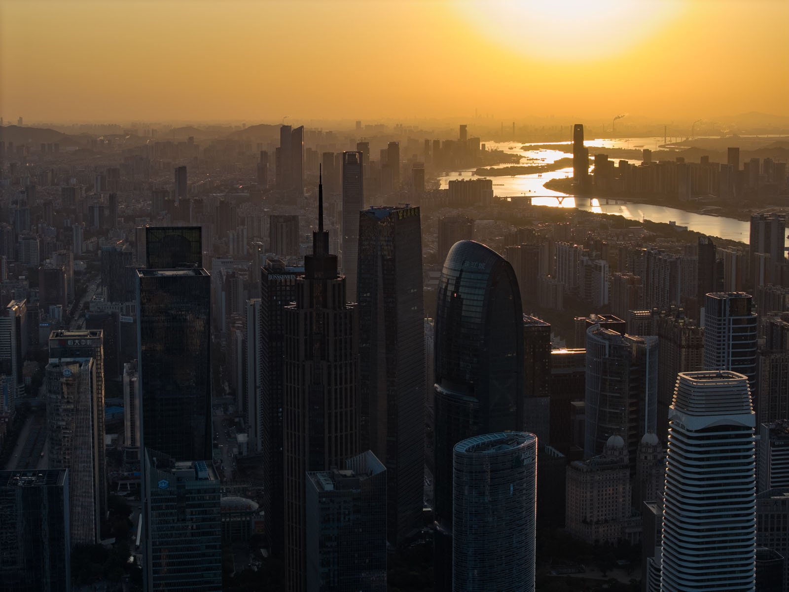 A city skyline at sunset with tall modern skyscrapers and a river winding through the buildings, all bathed in a warm golden light.