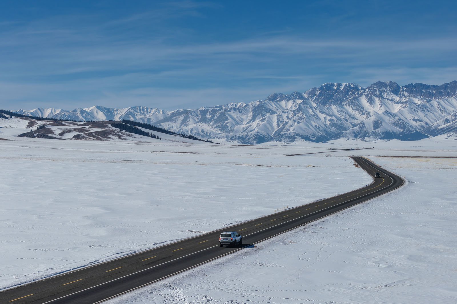A car drives along a winding road through a vast snowy landscape, with distant snow-covered mountains and a blue sky in the background.