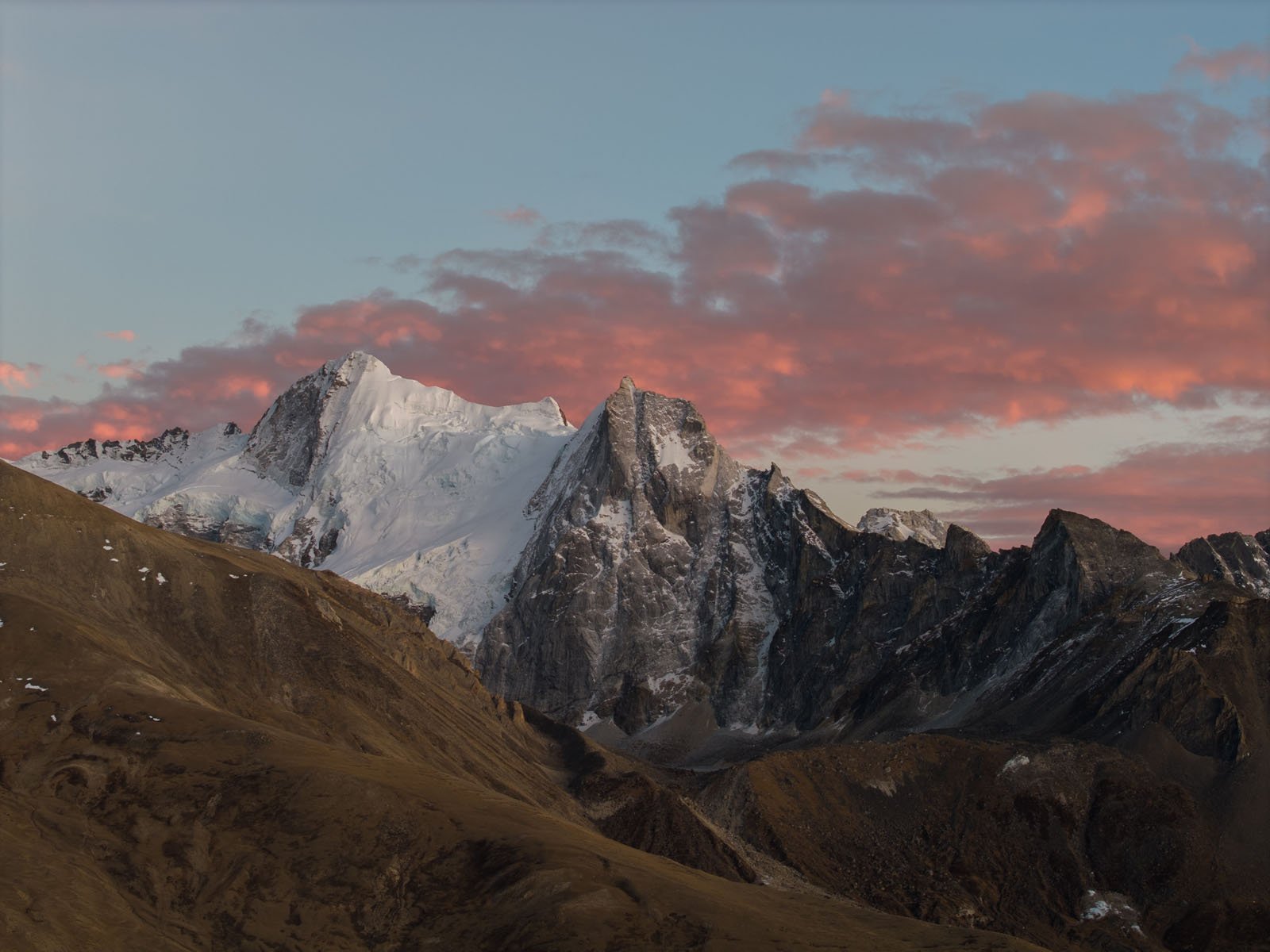 Snow-capped mountain peaks under a vibrant pink and orange sunset sky, with rugged brown slopes in the foreground and scattered clouds above.