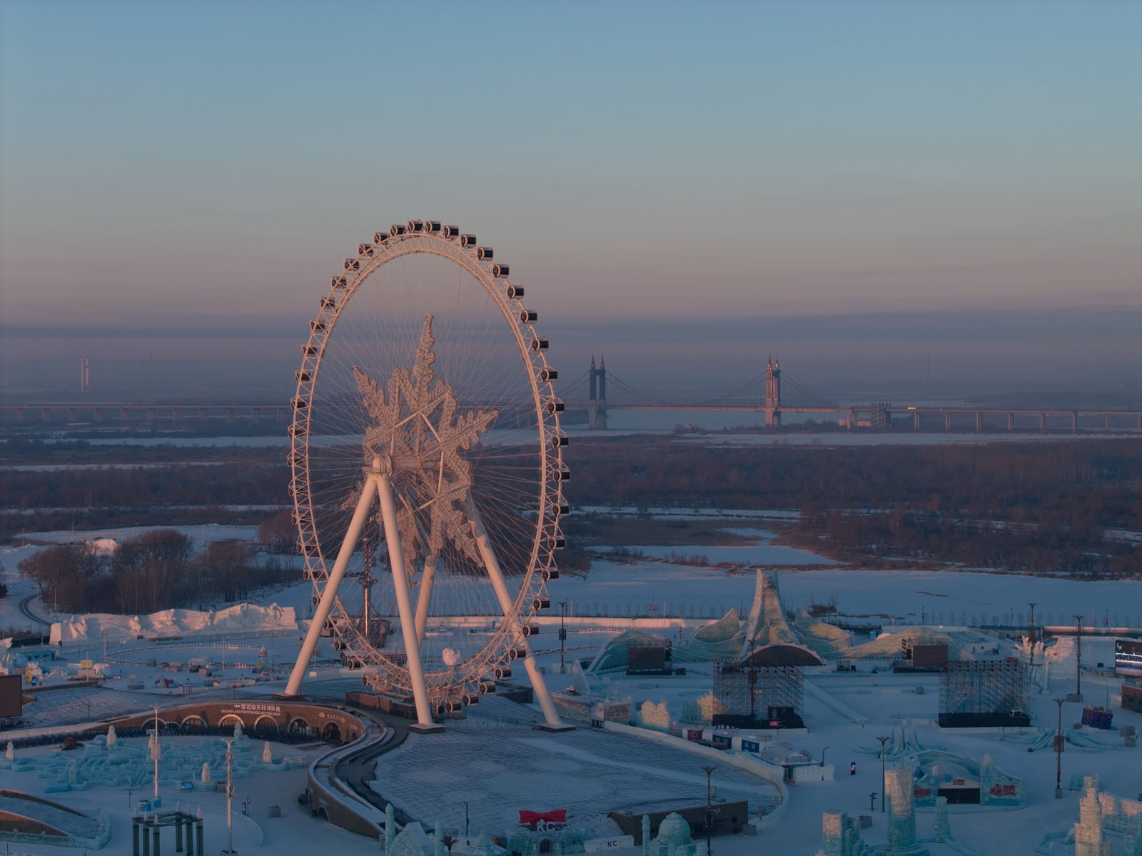 A large Ferris wheel decorated with a snowflake stands in a winter theme park, surrounded by ice and snow sculptures at dusk. A wide river and a bridge are visible in the background under a clear sky.