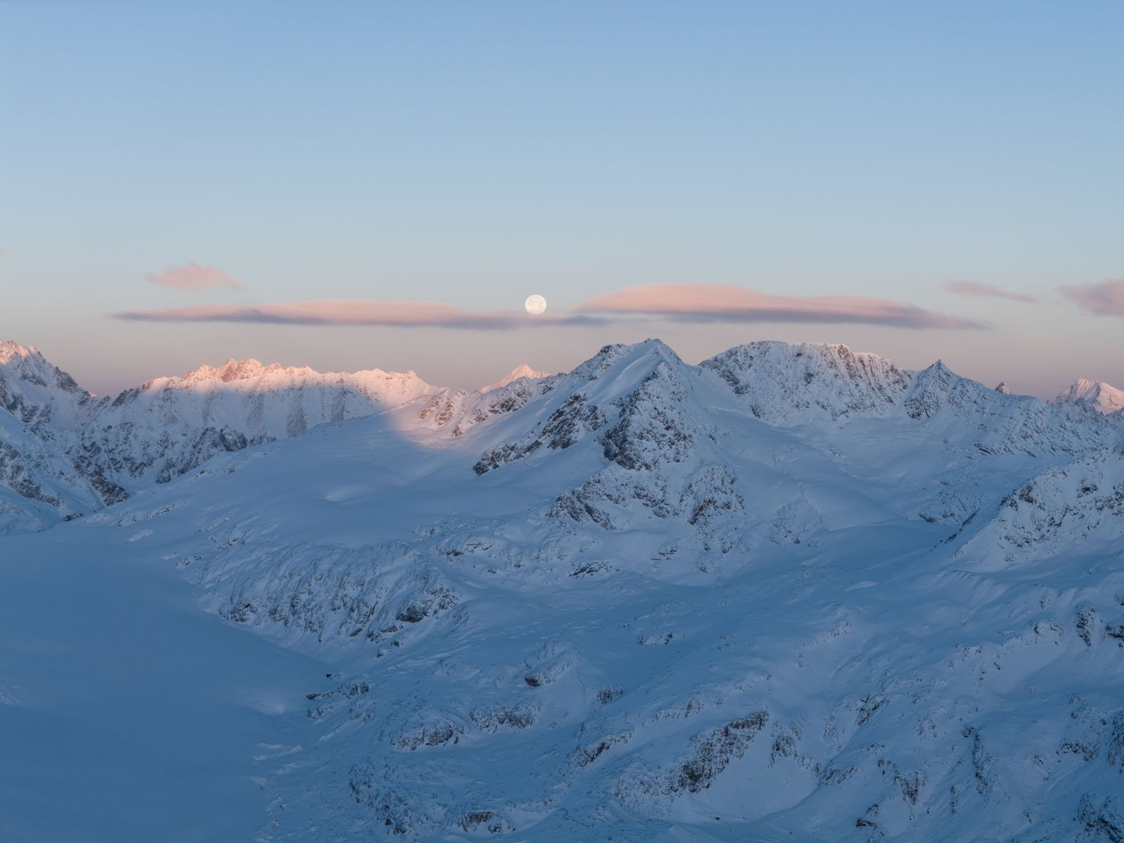 Snow-covered mountain peaks under a clear sky at dusk, with the full moon rising above the horizon and soft pink clouds illuminated by the setting sun.