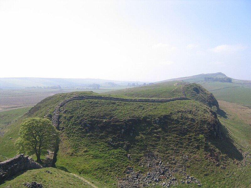 A grassy hill with the remains of an ancient stone wall stretching along the top, set in a rural landscape with open fields and distant rolling hills under a hazy sky.