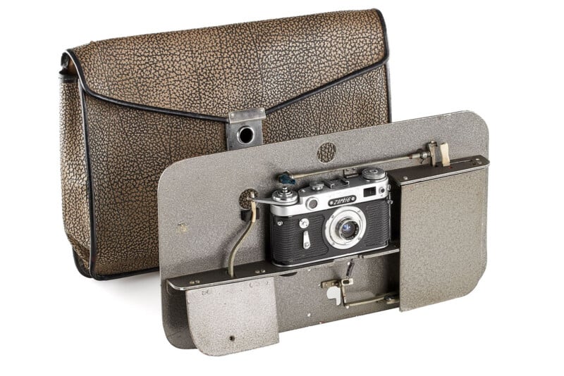 A vintage camera mounted on a flat metal plate with mechanical parts, displayed in front of a textured brown carrying case with a flap closure.