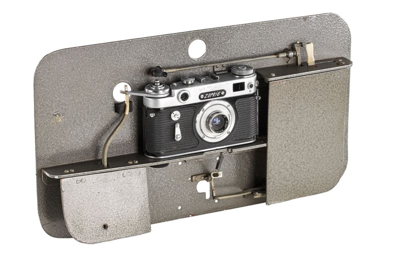 A vintage black and silver camera is mounted on a textured metal plate with mechanical components and levers beside it, possibly part of an old industrial or surveillance setup.