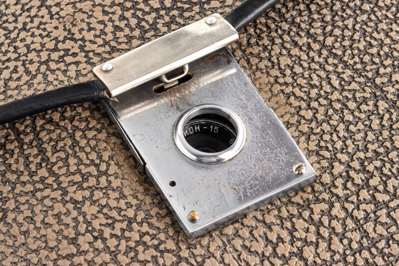 A close-up of a vintage metal camera shutter release mechanism attached to a textured, brown surface, with markings and a circular dial visible in the center.