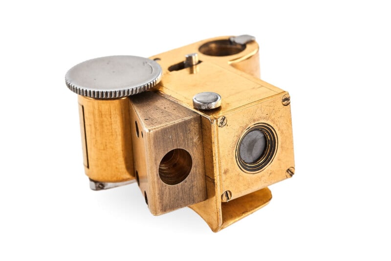 A small, vintage brass and metal camera with a boxy design, lens, and circular adjustment dial, photographed on a white background.