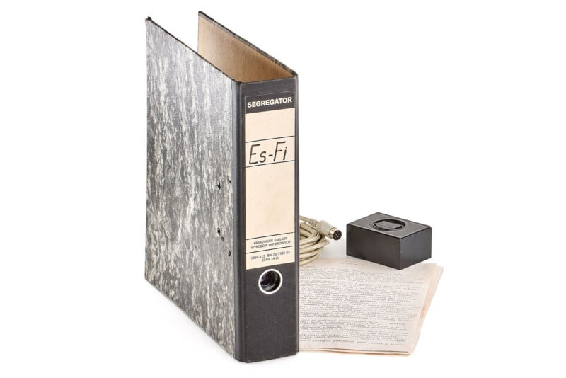 A gray marbled file binder labeled "Es-Fi" stands upright next to a magnifying glass, a metal key, and some scattered documents on a white background.
